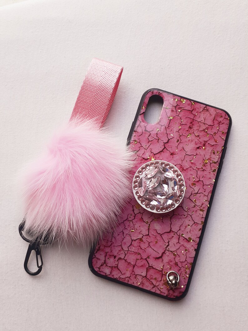 May include: A pink phone case with a cracked pink and gold design. The case has a clear rhinestone ring holder and a pink fluffy keychain with a silver clip.