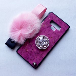 May include: A pink phone case with a glittery pink and gold design. The case has a clear phone grip with a rhinestone design. A pink fluffy pom-pom keychain is attached to the case.