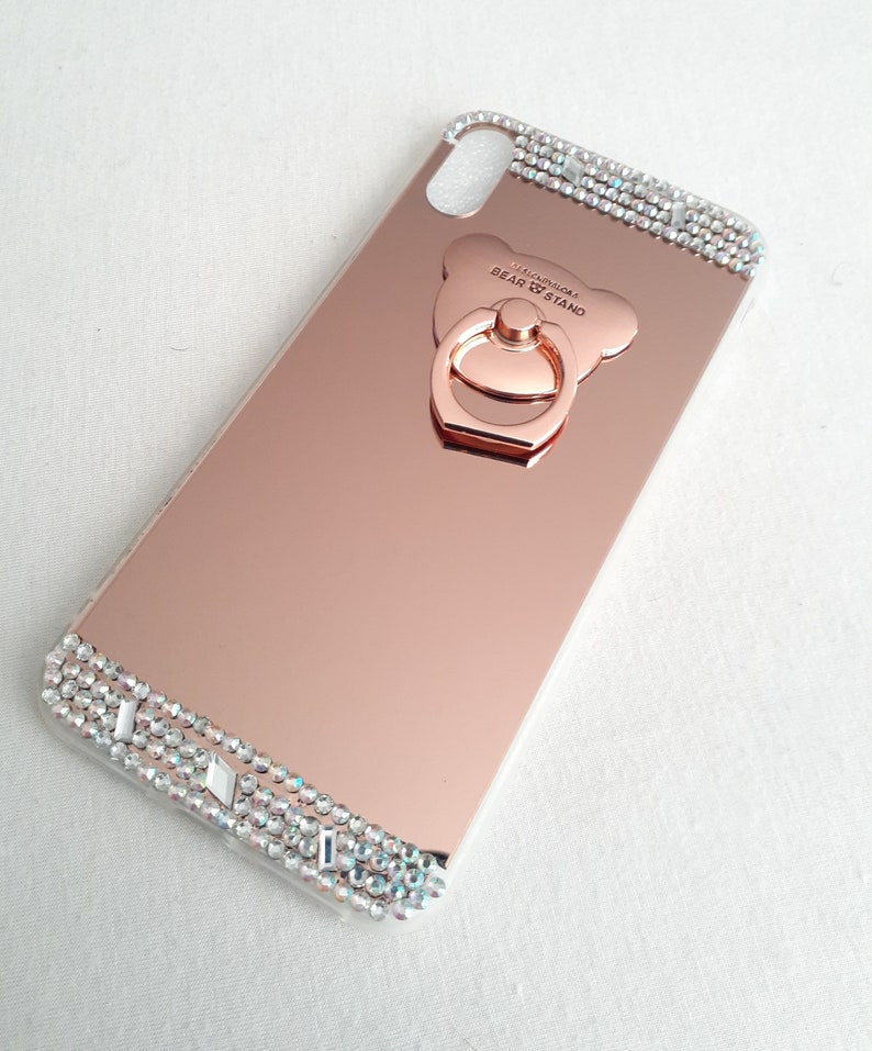 May include: Rose gold mirror phone case with a clear, sparkly, rhinestone trim and a rose gold bear-shaped ring holder with the text "FASHION BEAR STAND".