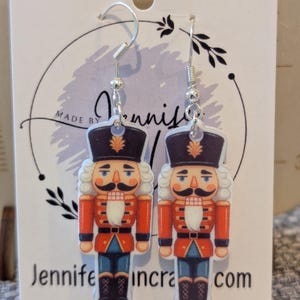 Könnte beinhalten: Ohrringe mit einem Nussknacker-Design. Die Ohrringe haben silberne Haken und zeigen eine detaillierte Illustration eines Nussknackersoldaten in roter Jacke, schwarzem Hut und blauer Hose. Auf der Karte steht "Jennifer incra.com" und "Sterling Silver."