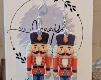 Puede incluir: Pendientes colgantes con diseño de cascanueces. Los pendientes tienen ganchos plateados y presentan una ilustración detallada de un soldado cascanueces con chaqueta roja, sombrero negro y pantalón azul. La tarjeta dice "Jennifer incra.com" y "Sterling Silver."