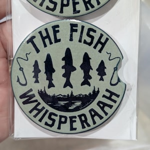 May include: Two round car coasters with a light green background. The text "THE FISH WHISPERAAH" is printed in dark blue, with fish and a fishing hook design. The coasters are packaged in clear plastic.