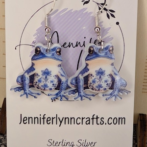 May include: A pair of frog-shaped earrings with a blue and white floral design, suspended from silver hooks. The earrings are displayed on a white card with the text "Jenniferlynncrafts.com" and "Sterling Silver".