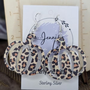 May include: A pair of pumpkin-shaped earrings with a leopard print pattern. The earrings are made of a light-colored material with brown and black spots. The earrings hang from silver-colored hooks. The text "Sterling Silver" is visible.