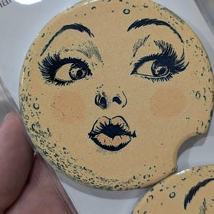 Puede incluir: Posavasos redondo de cer&aacute;mica beige con un rostro estilizado. El rostro tiene ojos, pesta&ntilde;as, cejas y labios azul oscuro. El posavasos tiene una peque&ntilde;a hendidura para un portavasos de coche. Peque&ntilde;os puntos oscuros.