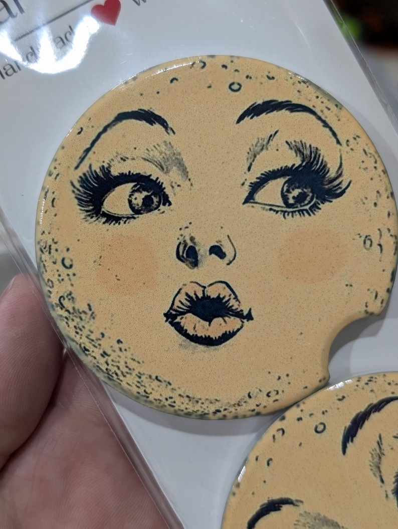 Puede incluir: Posavasos redondo de cer&aacute;mica beige con un rostro estilizado. El rostro tiene ojos, pesta&ntilde;as, cejas y labios azul oscuro. El posavasos tiene una peque&ntilde;a hendidura para un portavasos de coche. Peque&ntilde;os puntos oscuros.