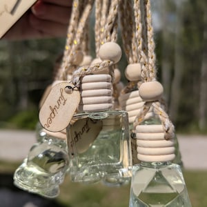 May include: A collection of clear glass aromatherapy bottles with wooden tops and 'Juniper' tags suspended from a braided rope. The bottles contain a pale green liquid. The rope has gold detailing. The background is out of focus.