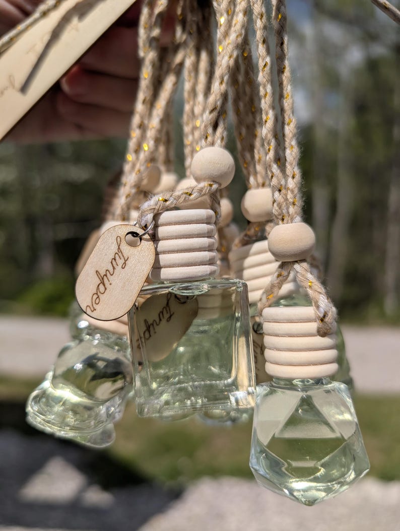 May include: Clear glass aromatherapy bottles with wooden tops and Juniper tags hang from a braided rope. The bottles are filled with a light green liquid. The rope has gold accents. The background is blurred.