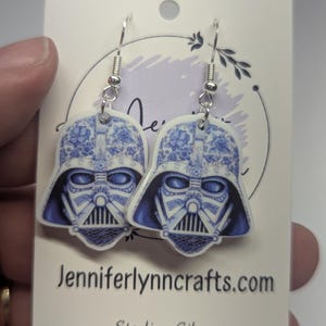 May include: A pair of blue and white patterned Darth Vader earrings with sterling silver hooks. The earrings are displayed on a white card with the text "jenniferlynncrafts.com" and "Sterling Silver".