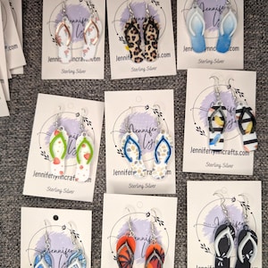 May include: A display of handmade sterling silver flip-flop earrings in various designs. Styles include leopard print, blue ombre, and beach-themed patterns. Each pair is displayed on a card with the text "Jennifer Lynn Crafts.com".