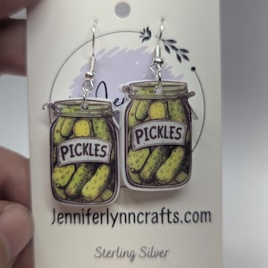 Pode incluir: Um par de brincos de prata esterlina com a forma de frascos de picles com a palavra "Pickles" no rótulo. Os frascos estão cheios de picles verdes.