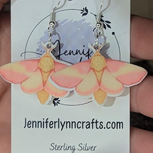 May include: A pair of moth-shaped earrings with pink and yellow wings, hanging from sterling silver hooks. The earrings are displayed on a white card with the text "Jenniferlynncrafts.com" and "Sterling Silver".