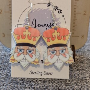 Può includere: Orecchini pendenti con un design a schiaccianoci a cartoni animati. Gli orecchini hanno ganci argentati e una carta bianca con la scritta "Sterling Silver". Lo schiaccianoci ha una corona, barba bianca e occhi blu.