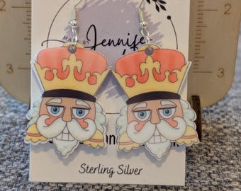 Puede incluir: Pendientes colgantes con un diseño de cascanueces de dibujos animados. Los pendientes tienen ganchos plateados y una tarjeta blanca con el texto "Sterling Silver". El cascanueces tiene una corona, barba blanca y ojos azules.
