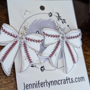 May include: A pair of baseball-themed bow earrings. The white bows feature red stitching details, mimicking a baseball. The earrings hang from silver hooks. The card behind the earrings reads "Jenniferlynncrafts.com" and "Sterling Silver".