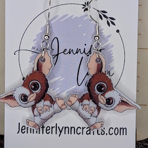 May include: A pair of sterling silver Gremlin earrings. Each earring features a cartoon Gremlin with brown, white, and tan coloring. The earrings are attached to silver fish hook ear wires. The backing card has the text "Jenniferlynncrafs.com" and "Sterling Silver".