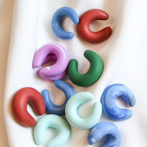 Könnte beinhalten: Eine Sammlung farbenfroher, C-förmiger Keramikobjekte in verschiedenen Blau-, Rot-, Grün- und Lilatönen. Die Objekte sind auf einem weißen Stoffhintergrund angeordnet und zeigen ihre gebogenen Formen und bemalten Oberflächen.