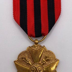 May include: A gold-coloured medal with a starburst design and a red and black striped ribbon. The medal has an ornate, detailed design with a central emblem. The ribbon is attached to the medal with a gold ring.