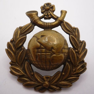Peut inclure: Un emblème de couleur bronze représentant un globe terrestre au centre, entouré d'une couronne de laurier. Au-dessus du globe, deux cornes incurvées et un nœud décoratif. L'emblème a un aspect antique détaillé.
