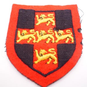 Puede incluir: Parche bordado rojo y negro con forma de escudo. El parche presenta un borde rojo, un diseño de cruz negra y leones bordados en amarillo. Probablemente sea un emblema militar o heráldico.