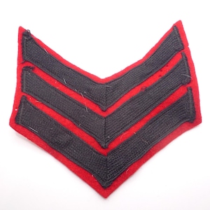 May include: A red and black chevron-shaped patch. The patch has a red border and three black chevrons. The patch is likely a military or service rank insignia.