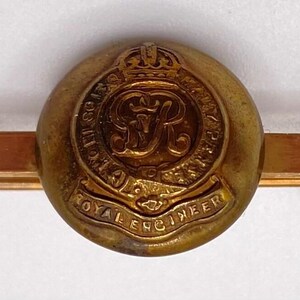 Puede incluir: Un pasador de corbata dorado con un emblema circular con una corona y el texto "ROYAL ENGINEER". El pasador tiene una barra rectangular y un extremo curvado para sujetarlo a la corbata. El emblema tiene las letras "RR" en el centro.