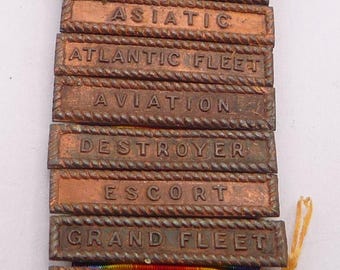 May include: A collection of vintage military service ribbons and bars. The bronze bars are engraved with phrases like "ARMED GUARD" and "OVERSEAS." The ribbons display a rainbow color scheme.