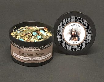Puede incluir: Una lata de vela negra con el texto "Egyptian Deities" y "In Honor of Ma'at". La vela está llena de hierbas secas y piedras turquesas. La tapa presenta una ilustración de una mujer con un tocado. Peso neto 200 g.