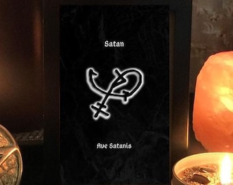 May include: A framed black print with the word "Satan" at the top and "Ave Satanis" at the bottom. A white symbol is in the center. A lit candle and a salt lamp are in the background.