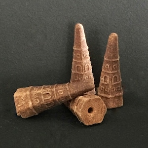 May include: Three brown, cone-shaped incense items with a textured, architectural surface pattern featuring small arched windows. One cone is lying horizontally, while two others stand vertically, showing a circular hole at the base.