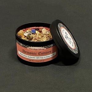 May include: A black metal candle tin with a peach-colored label reading Silver Moon Offerings Demonic Entities Researched, Authentic Offerings in Every Candle, Net Wt. 7.4 oz (209 g). The candle is topped with various small, colorful stones.