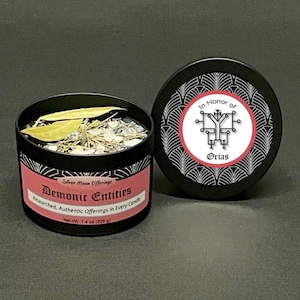 May include: Black candle tin with the text "Demonic Entities" and "Silver Moon Offerings". The candle is filled with dried herbs and crystals. The lid features a sigil and the words "In Honor of Orias". Net weight is 7.4 oz (209 g).