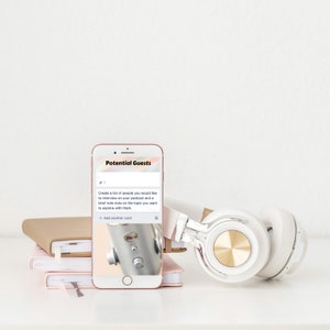 May include: A rose gold smartphone displays the text "Potential Guests" and a list of podcast interview ideas. White and gold headphones rest beside the phone, with a stack of notebooks in the background. The image suggests a podcasting or content creation theme.