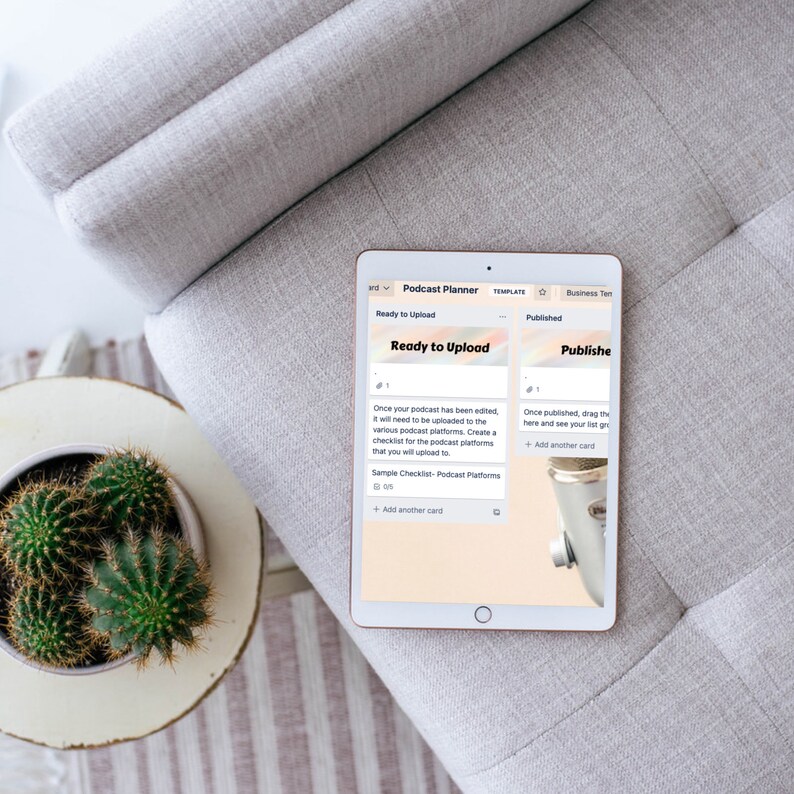 May include: A digital tablet displaying a podcast planner interface, with sections labeled "Ready to Upload" and "Published." The tablet rests on a gray upholstered chair. A small cactus in a white pot sits on a side table.