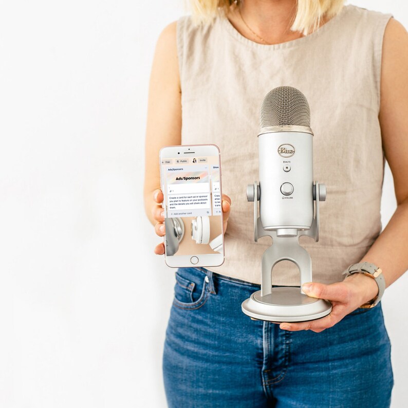 May include: A silver Blue Yeti USB microphone with a mesh top is held in front of a person. The person also holds a smartphone displaying a social media ad. The microphone has a silver stand and control buttons. The phone shows a picture of headphones.