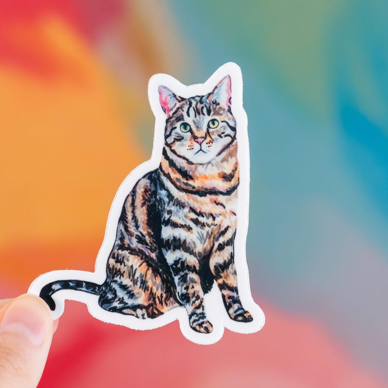 May include: A sticker featuring a watercolor illustration of a tabby cat in a seated position. The cat has brown and black stripes, green eyes, and pink ears. The sticker is held by a hand against a colorful, blurred background.