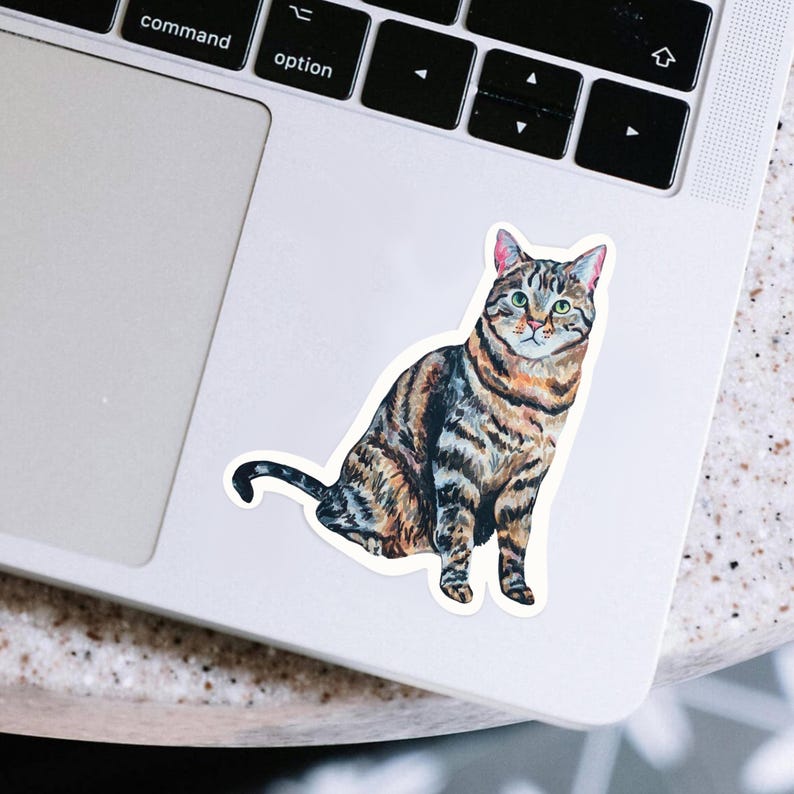 May include: A sticker featuring a watercolor illustration of a tabby cat in shades of brown, black, and orange. The cat is sitting upright with its tail curled. The sticker is placed on a white laptop.