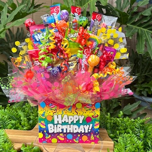 May include: A colorful candy bouquet in a box with the words "Happy Birthday!" The bouquet includes various candies, lollipops, and ribbons. The box is decorated with a festive pattern of party hats and balloons. The arrangement is set on a wooden surface.