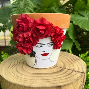 May include: A white ceramic plant pot with a hand-painted portrait of Frida Kahlo, red lips, and a floral crown. The pot has a terracotta rim and is decorated with red fabric flowers. The pot is displayed on a wooden stand.