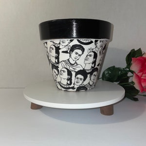 Puede incluir: Una maceta de cerámica en blanco y negro con un patrón repetido del retrato de Frida Kahlo. El borde de la maceta está pintado de negro. La maceta se exhibe sobre un soporte redondo con patas de madera blancas.