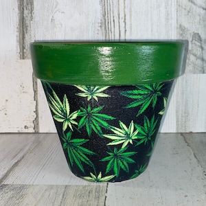 Può includere: Un vaso di fiori in terracotta dipinto con bordo verde e base nera. La base è decorata con un motivo di foglie di cannabis verdi. Per piantare fiori o erbe.