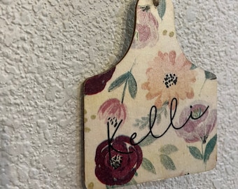 May include: A floral patterned wooden ornament with the name "Kelli" written in cursive. The ornament is attached to a twine string with two wooden beads.