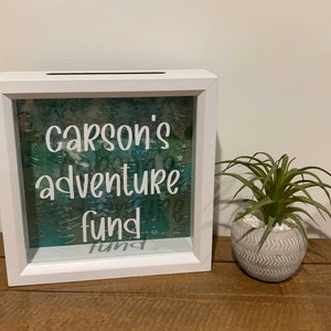 May include: A white shadow box with the text "Carson's adventure fund" in white script. The background features a teal and green forest design. A small potted plant with green leaves sits to the right.