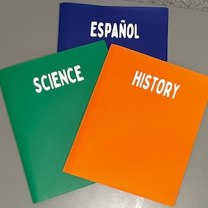 May include: Three plastic folders with different colored covers. The folders are labeled "Science" in green, "History" in orange, and "Español" in blue.