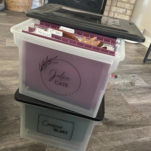May include: Two clear plastic storage boxes with black lids. The top box has a purple file folder with the name "Jolen Cate" written on it. The bottom box has a green file folder with the name "Cooper Blake" written on it.