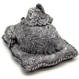 May include: Silver-colored figurine of a dog lying on a pillow with wings. The dog is sleeping with its head resting on the pillow. The figurine is made of a detailed, textured material.