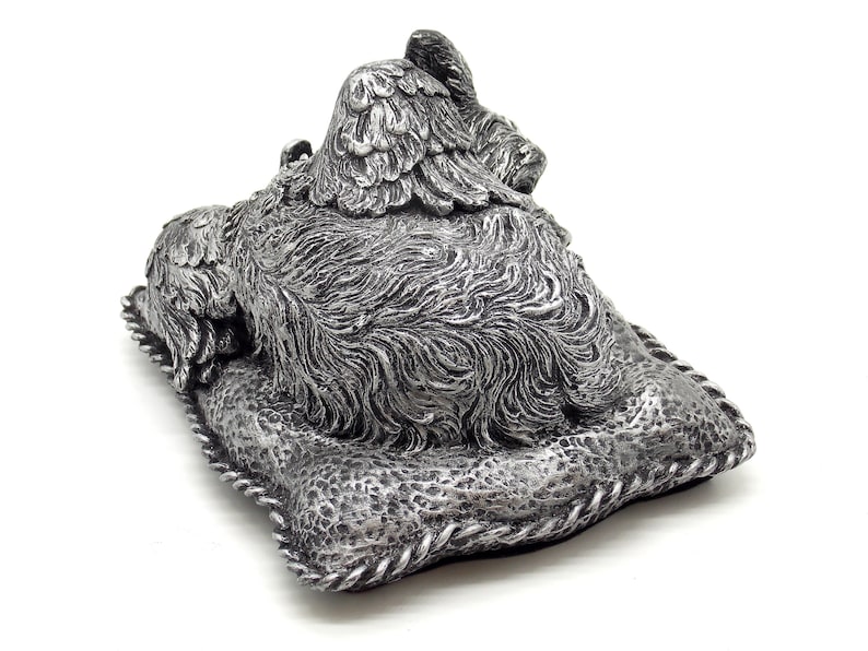 May include: Silver-colored figurine of a dog lying on a pillow with wings. The dog is sleeping with its head resting on the pillow. The figurine is made of a detailed, textured material.