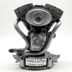 May include: A silver-colored motorcycle engine sculpture. The engine features detailed components, including cylinders, pipes, and a central housing. The base has a banner with the name "Dustin Lee Pope" and dates.