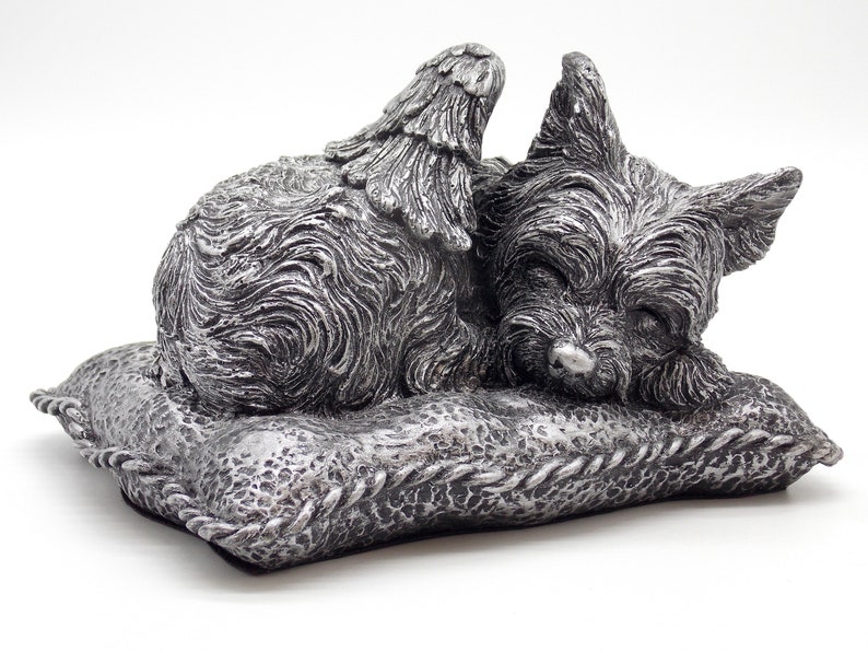 May include: Silver-colored figurine of a sleeping Yorkshire Terrier with angel wings, resting on a pillow. The figurine is a pet memorial.