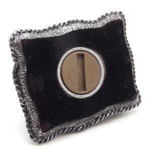 May include: A silver belt buckle with a black velvet inlay and a silver-toned metal insert. The buckle has a rope-like design around the edge.